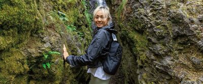 A woman stands between two mossy rocks on a hiking trail looking at the camera.  